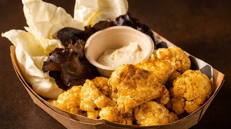 Crispy fried cauliflower bites with a side of creamy dip and fresh lettuce in a paper tray.
