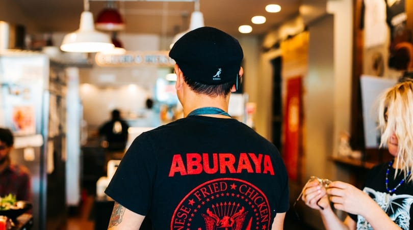 Back of a person wearing a black "Aburaya Japanese Fried Chicken" t-shirt in a restaurant.