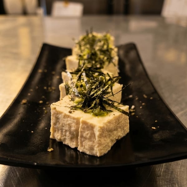 Three pieces of tofu topped with green sauce and seaweed on a black rectangular plate.