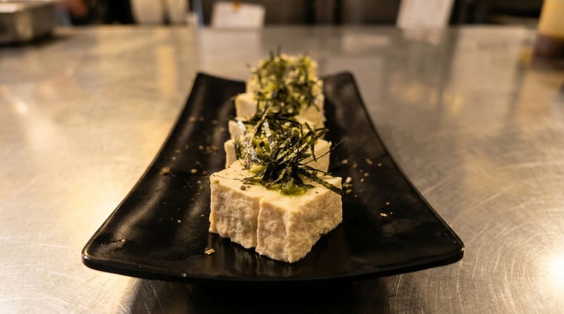Three pieces of tofu topped with green sauce and seaweed on a black rectangular plate.