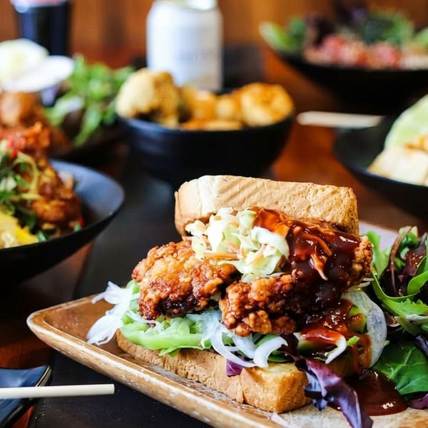 A fried chicken sandwich with coleslaw and BBQ sauce, with various Asian dishes in the background.