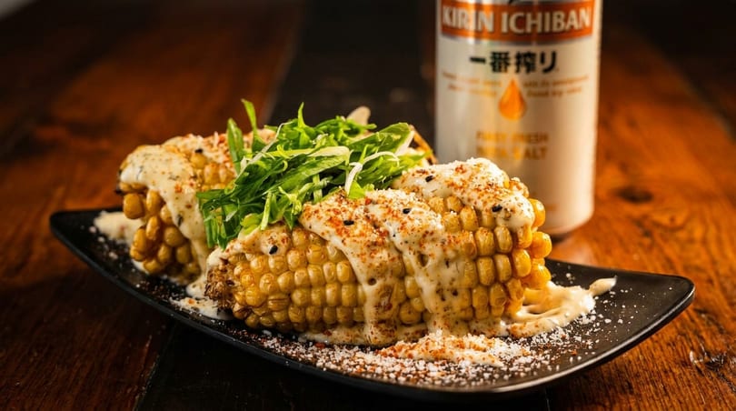 Grilled corn on the cob with creamy sauce, spices, and greens, next to a Kirin Ichiban can.