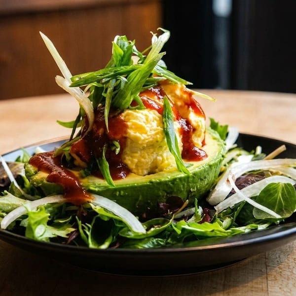 Avocado half filled with yellow dip, topped with green onions and red sauce, on a bed of greens.