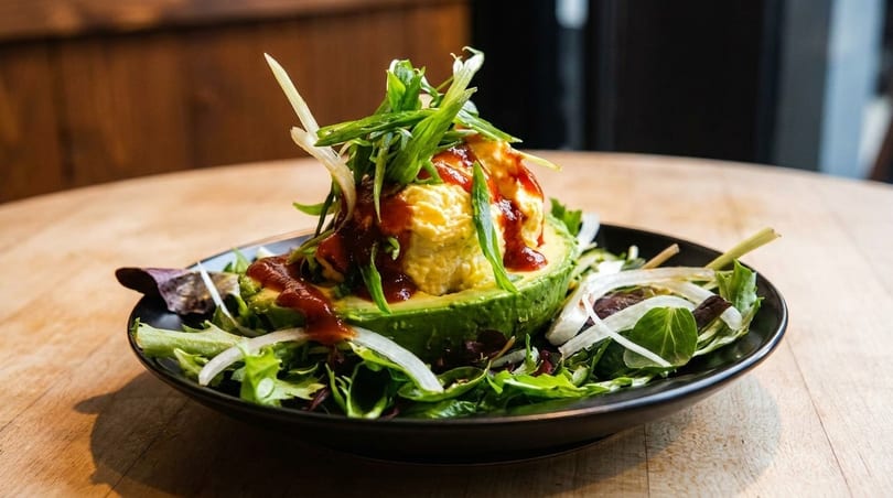 Avocado half filled with yellow dip, topped with green onions and red sauce, on a bed of greens.